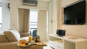 Quarto de hotel moderno com sofá, mesa de jantar com café da manhã, televisão e uma janela grande.
