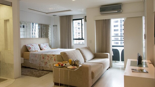 Quarto de hotel com cama, sofá, pequena mesa com itens de café da manhã, TV e janela com vista para a cidade.
