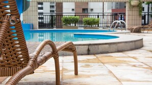 Área de piscina com cadeiras de vime, piso ladrilhado e um edifício ao fundo.