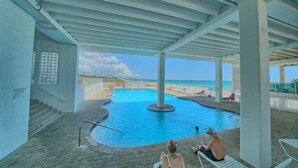 Una piscina cubierta con vista a la playa, una escalera que lleva a un nivel superior y algunas personas relajándose junto a la piscina.