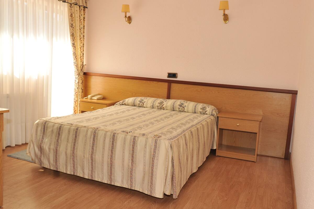 A hotel room with a wooden bed, a nightstand, a bedside table, and a wall-mounted lamp.