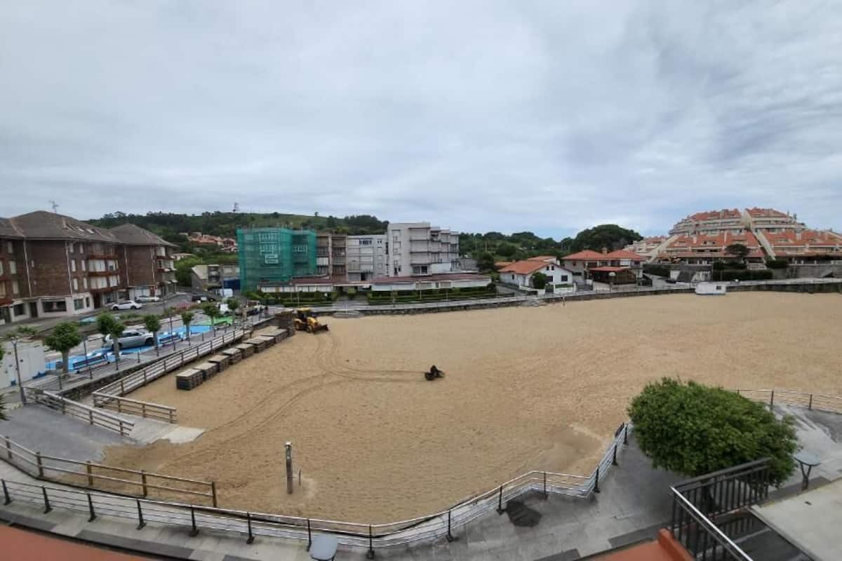 A large sandy sports field with a fenced perimeter, surrounded by buildings and a hill in the background.