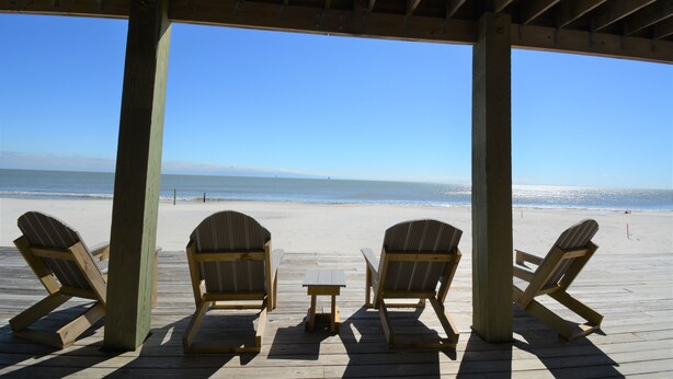 A beachside wooden deck with three Adirondack chairs and a small table, overlooking the ocean.