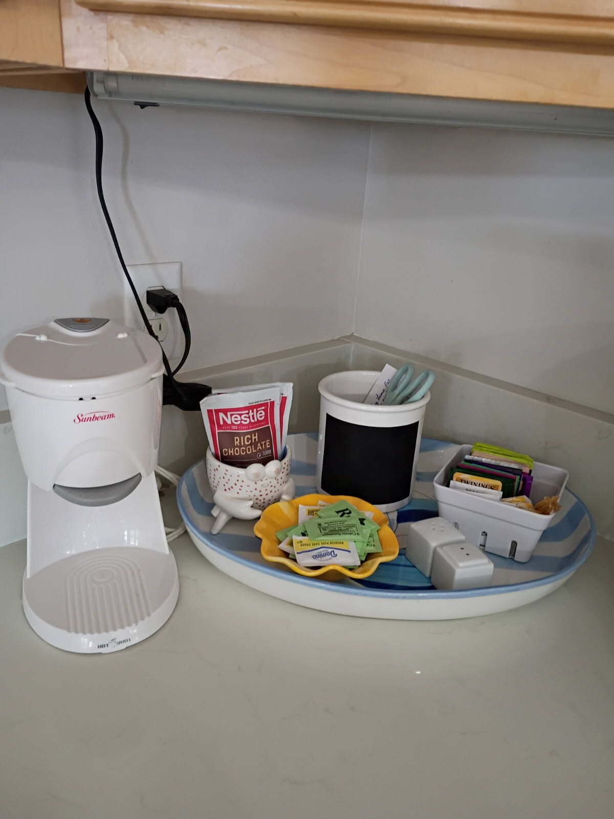 A kitchen area with a coffee maker, a coffee pot, a cup of Nestle Rich Chocolate, a bowl of money, and a small trash bin.