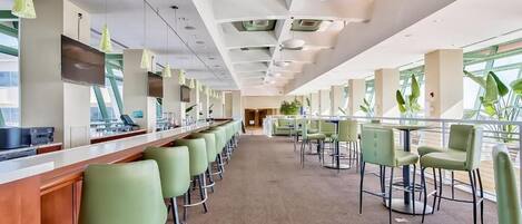 A modern bar area with green bar stools, a long counter, and large windows providing natural light.