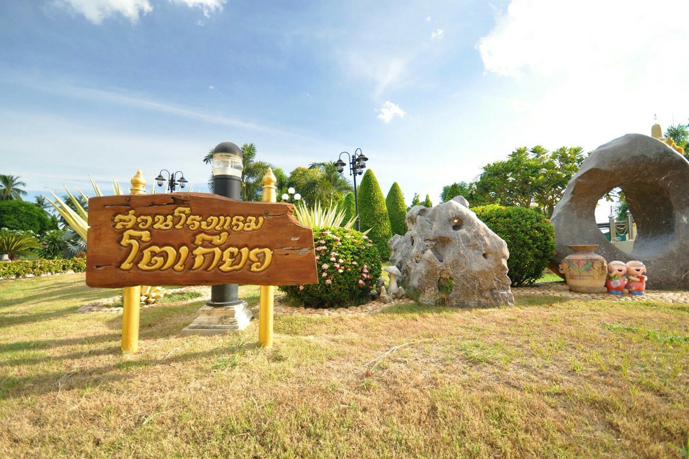 A wooden sign with Thai script, a stone sculpture, and a large rock formation.