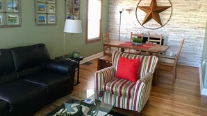 Ein Wohnzimmer mit einer Ledersofa, einem gestreiften Sessel mit rotem Kissen, einem Glastisch, einem Holzesstzimmer mit Stühlen, einer Stern-Dekoration und gerahmten Bildern an der Wand.