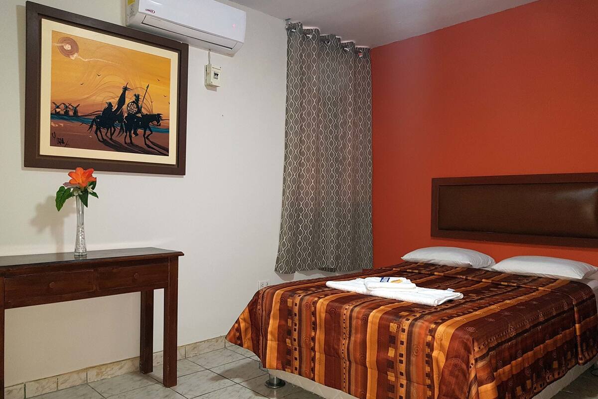 Ett hotellrum med en säng, ett skrivbord, en inramad tavla, en luftkonditionering och en gardin.