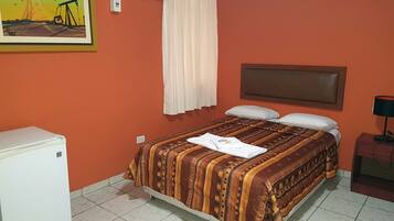 A hotel room with a bed, a small refrigerator, an air conditioner, a nightstand, and a painting on the wall.