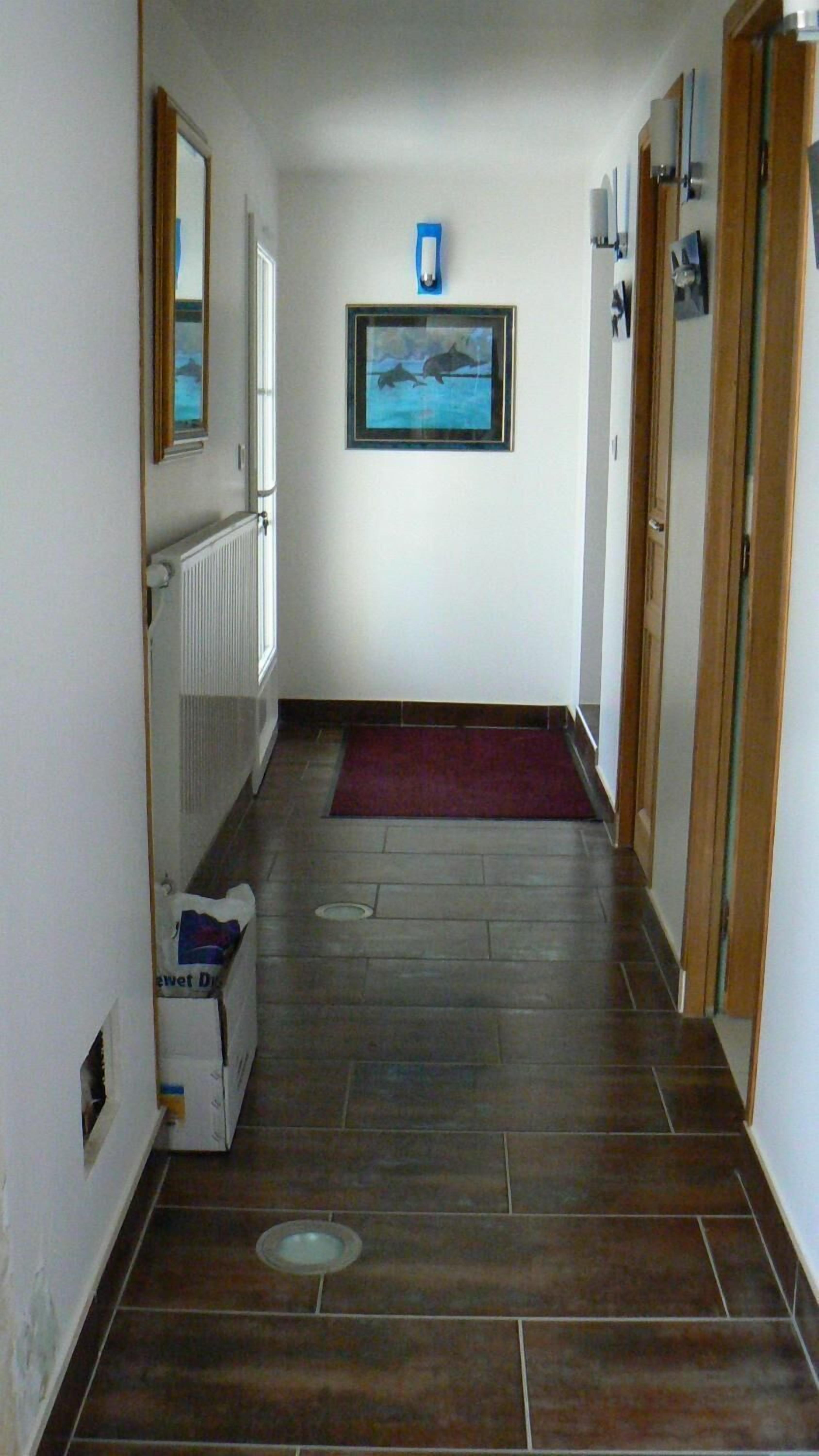 Un couloir étroit au sol carrelé, avec un tableau encadré au mur et un radiateur sur le côté gauche.