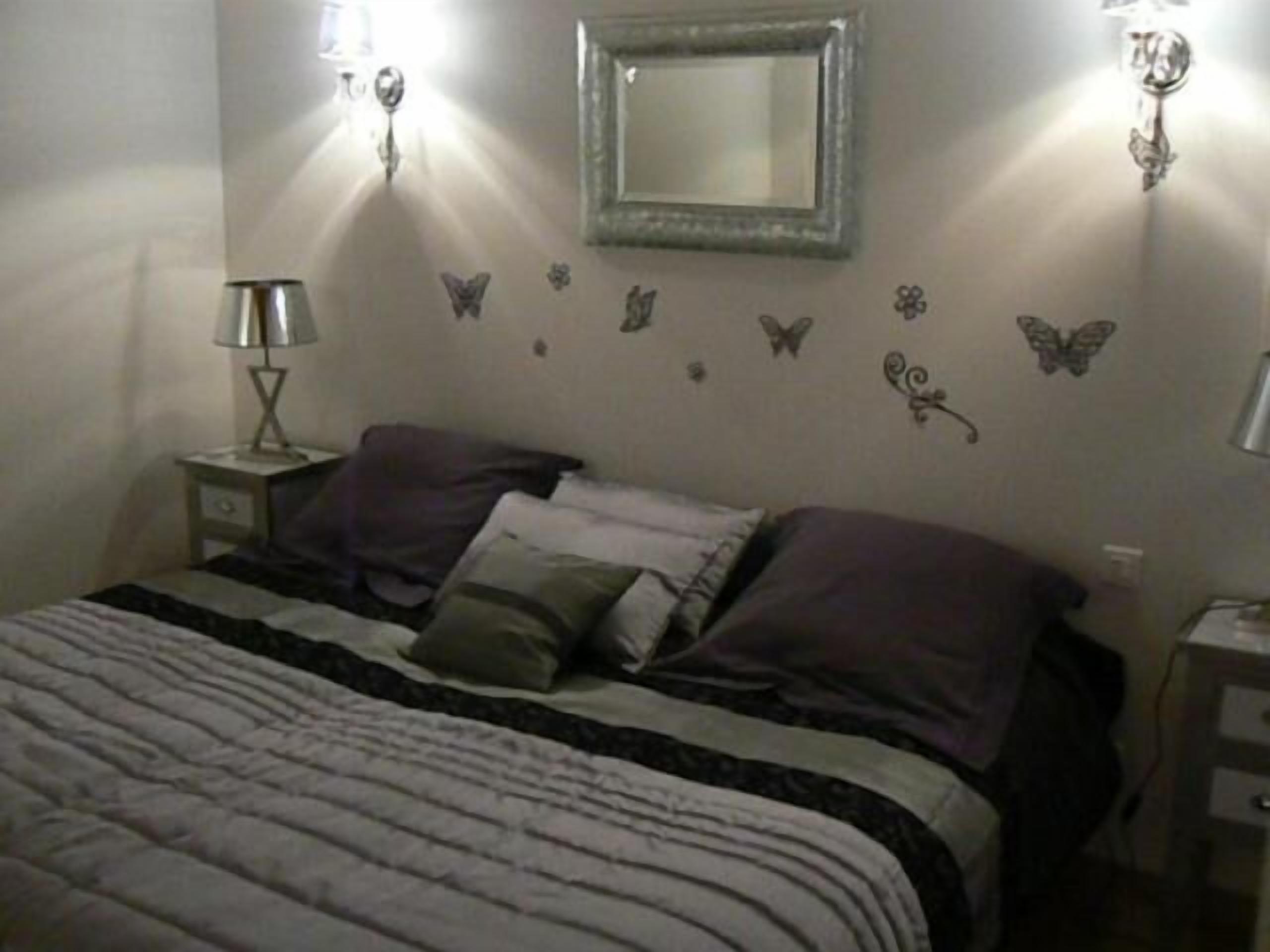 Een slaapkamer met een bed, kussens, een nachtkastje, een lamp, een spiegel en wanddecoraties, waaronder vlinders en bloemen.