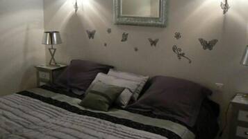 Ein Schlafzimmer mit Bett, Kissen, einem Nachttisch, einer Lampe, einem Spiegel und Wanddekorationen, darunter Schmetterlinge und Blumen.