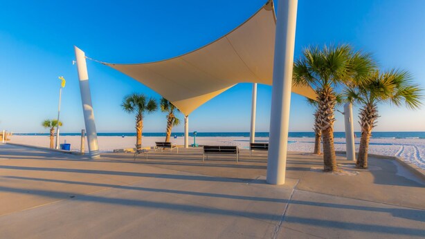 Une promenade en bord de mer bordée de palmiers, agrémentée de bancs et surmontée d’une grande structure en forme de tonnelle.
