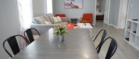 Un comedor con una mesa, sillas y un jarrón con plantas. Se observa un sofá y un cuadro en la pared al fondo.