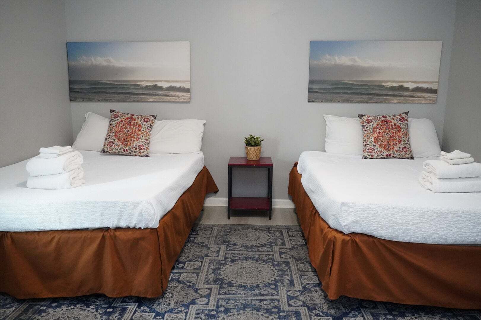 Ein Hotelzimmer mit zwei Betten, jeweils mit weißer Bettwäsche und braunen Tagesdecken, einem kleinen Nachttisch mit einer Pflanze sowie zwei an der Wand hängenden Bildern einer Strandlandschaft.