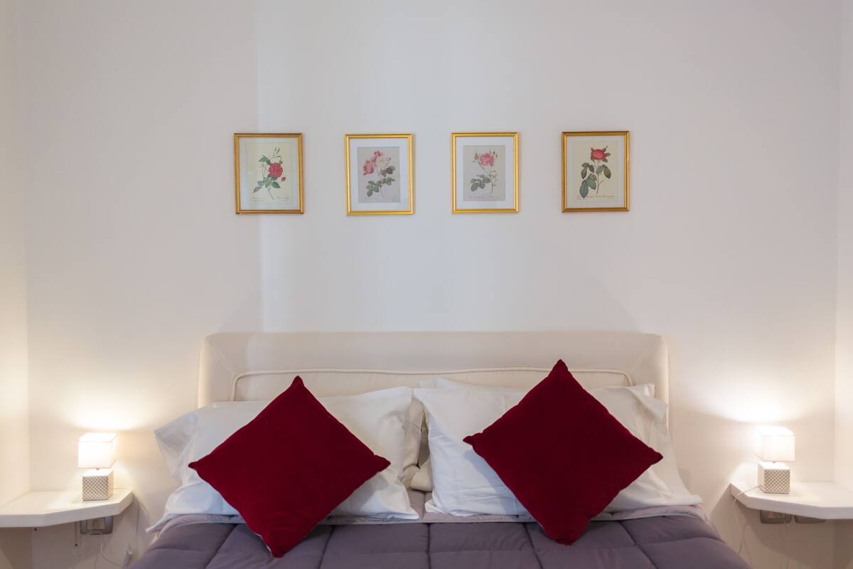 Ein ordentlich bezogenes Bett mit roten Kissen, einem weißen Kopfteil und vier gerahmten Pflanzenbildern an der Wand.