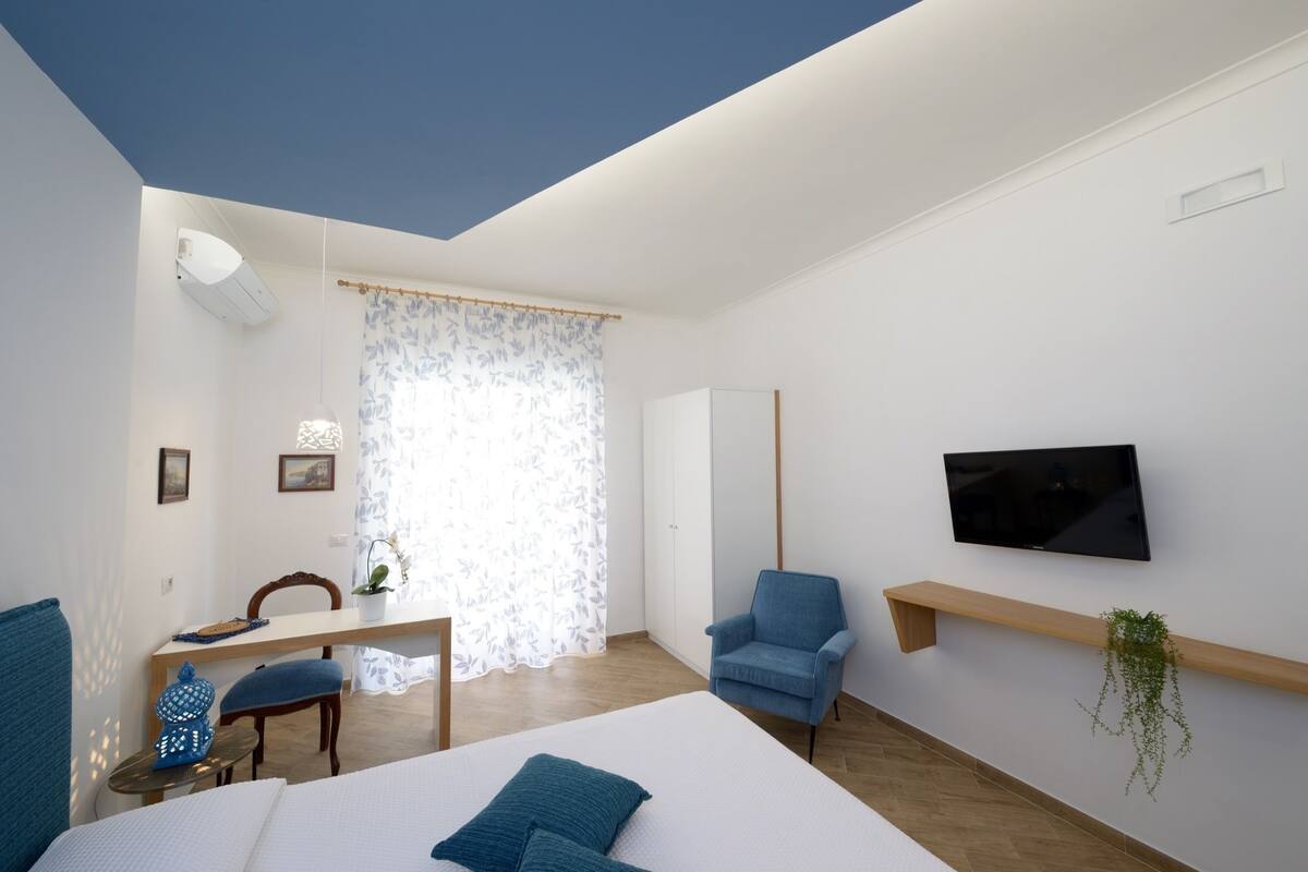 Ein modernes Schlafzimmer mit Bett, Schreibtisch, Stuhl, Fernseher und einem Fenster mit Vorhängen.