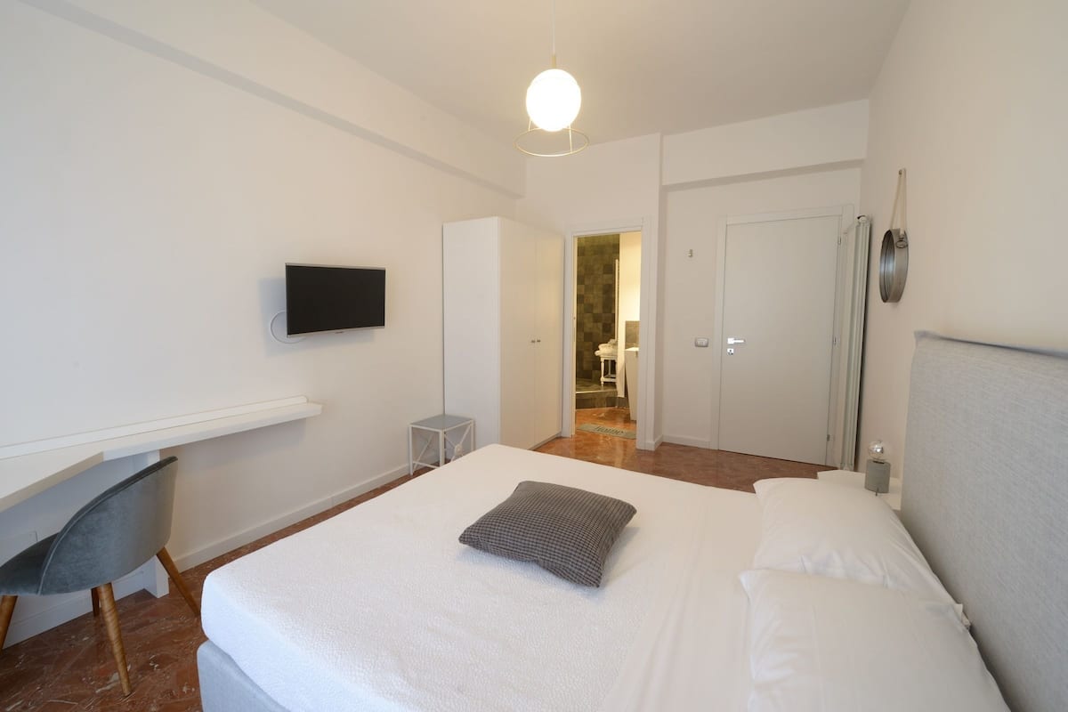 Ein modernes Hotelzimmer mit einem großen Bett, einem flachbildfernseher an der Wand, einem kleinen Schreibtisch und einem Sessel.