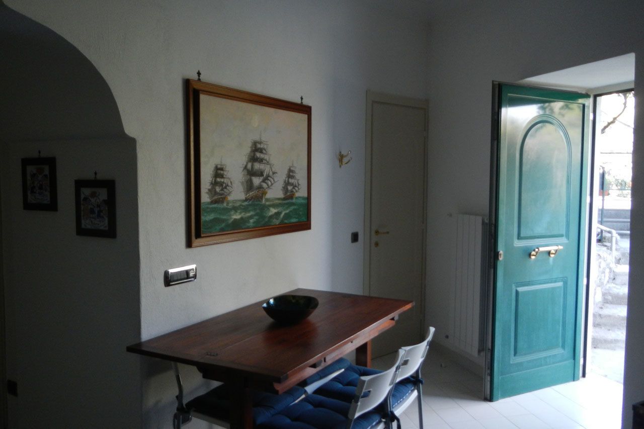 Ein Essbereich mit einem Holztisch, blau gepolsterten Stühlen, einem an der Wand hängenden Gemälde von Schiffen und einer türkisblauen Tür.