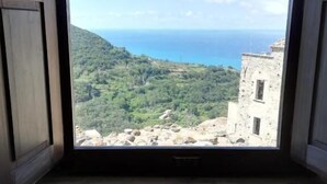 A room with a large window offering a view of a coastal landscape, including a stone building and greenery. - A Taverna Intru u vicu (Belmonte Calabro)