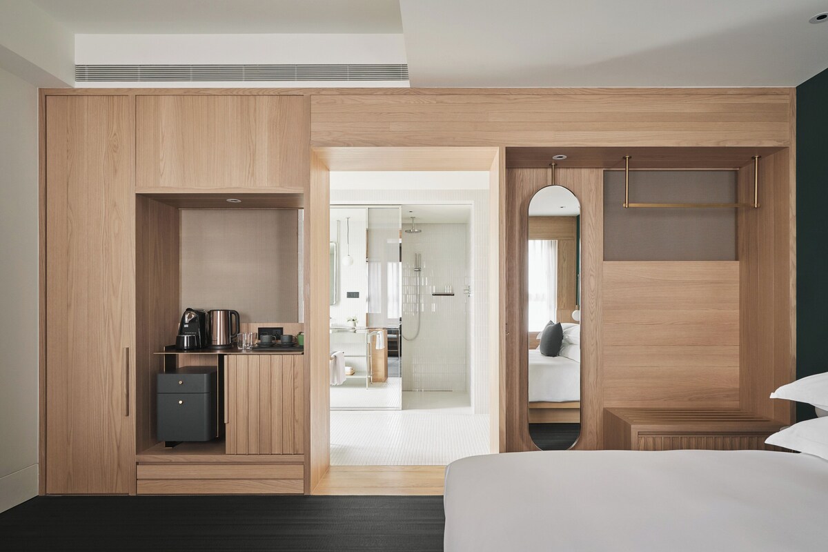 Une chambre d’hôtel moderne avec une armoire en bois, un petit bureau avec une bouilloire, un lit et un miroir.
