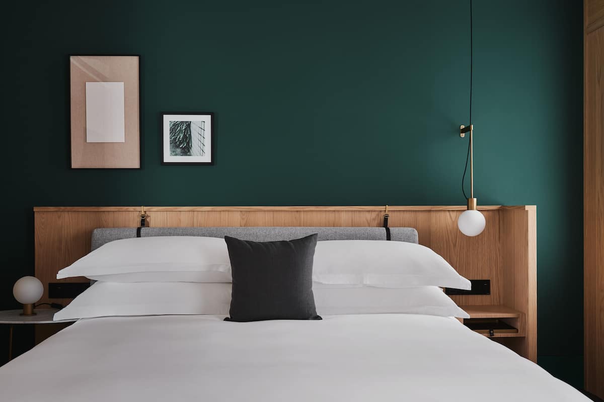 Un lit bien fait, avec une literie blanche et grise, une tête de lit en bois et une table de chevet sur laquelle se trouve une lampe.