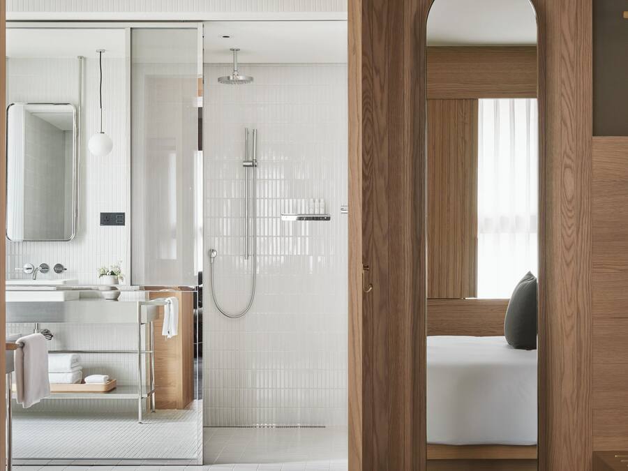 A modern bathroom with a shower, sink, and mirror. There is a wooden door leading to a bedroom with a bed and a window.