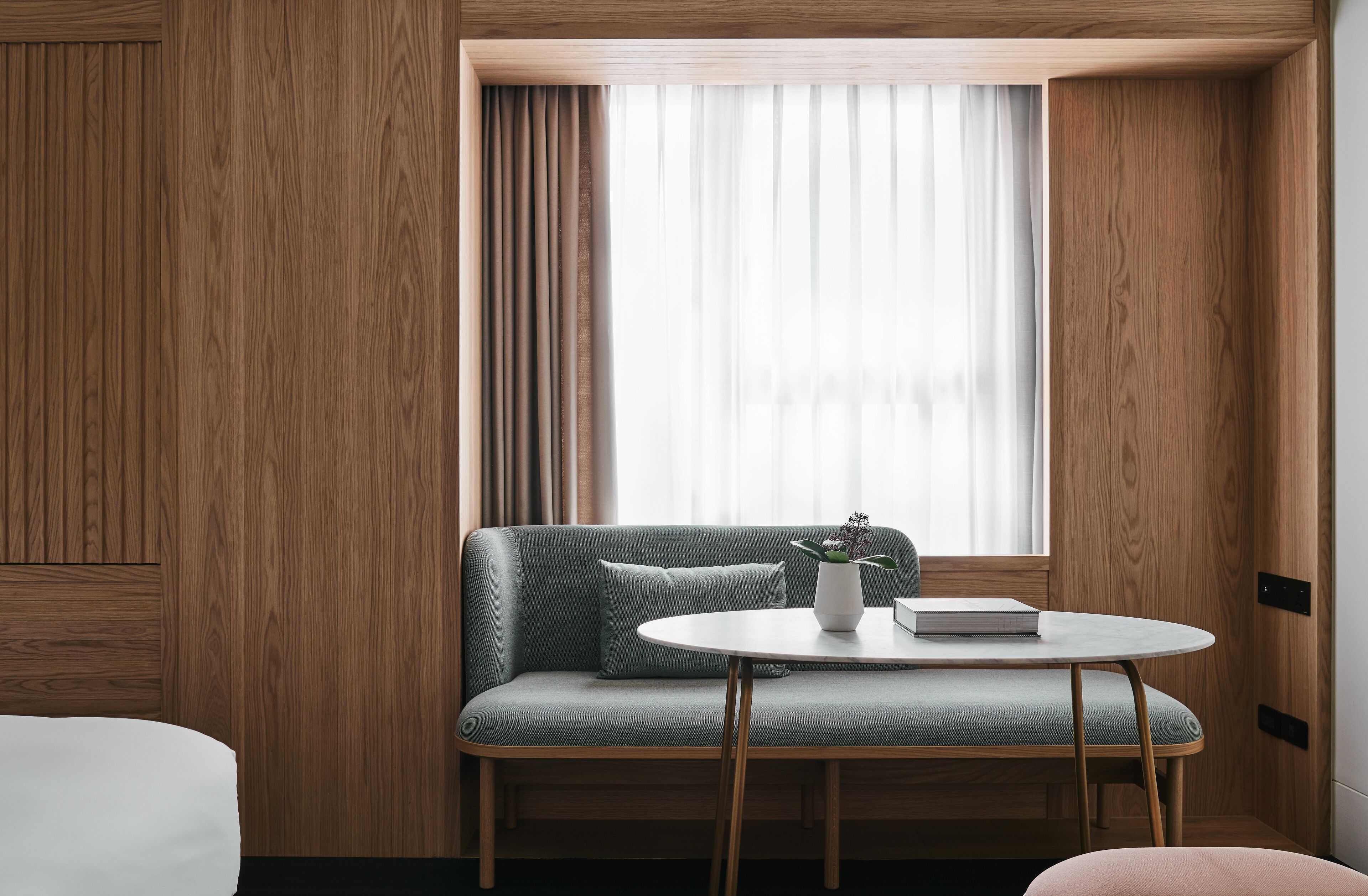 Una habitación de hotel moderna con pared de paneles de madera, un sofá gris, una pequeña mesa redonda con un libro y un florero, y una ventana con cortinas translúcidas.