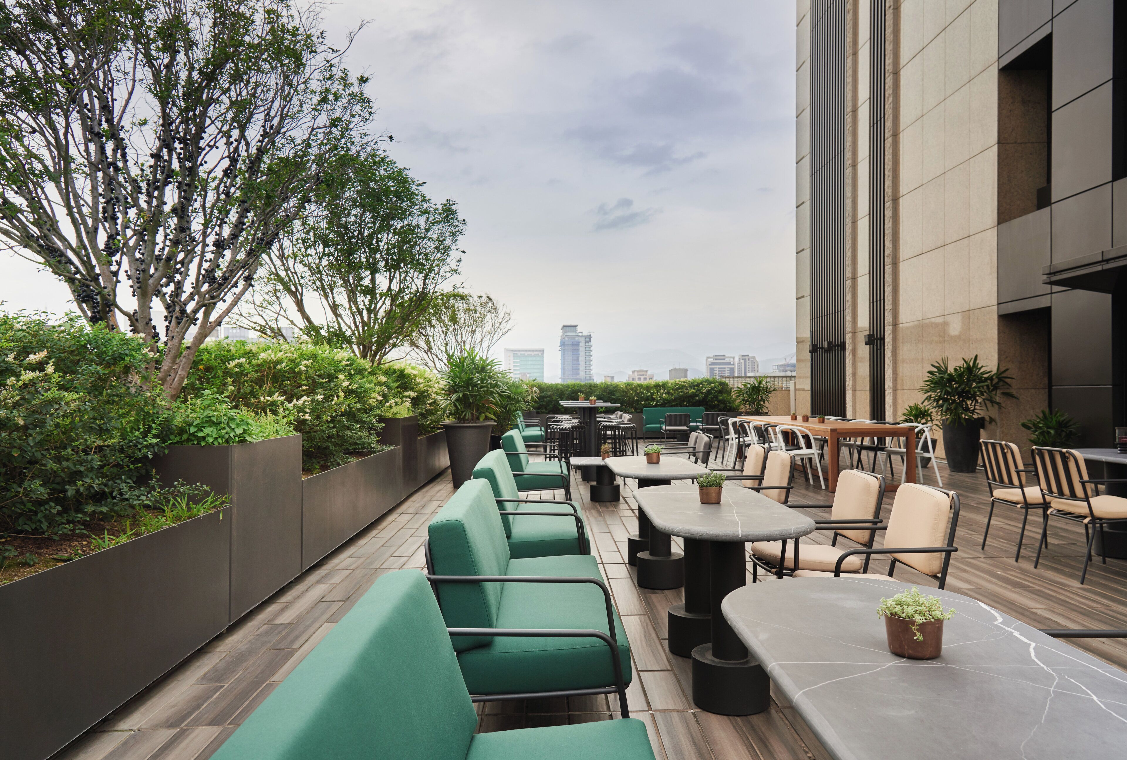 An outdoor seating area with green cushioned benches and tables, surrounded by greenery and a cityscape view.