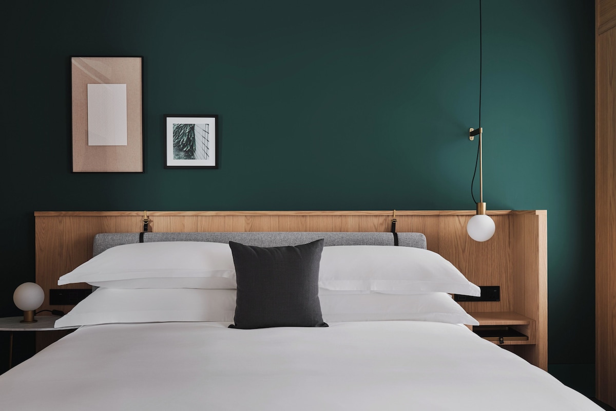 Une chambre moderne avec une tête de lit en bois, une literie blanche, un oreiller noir, une table de chevet avec une lampe et deux tableaux encadrés accrochés au mur.