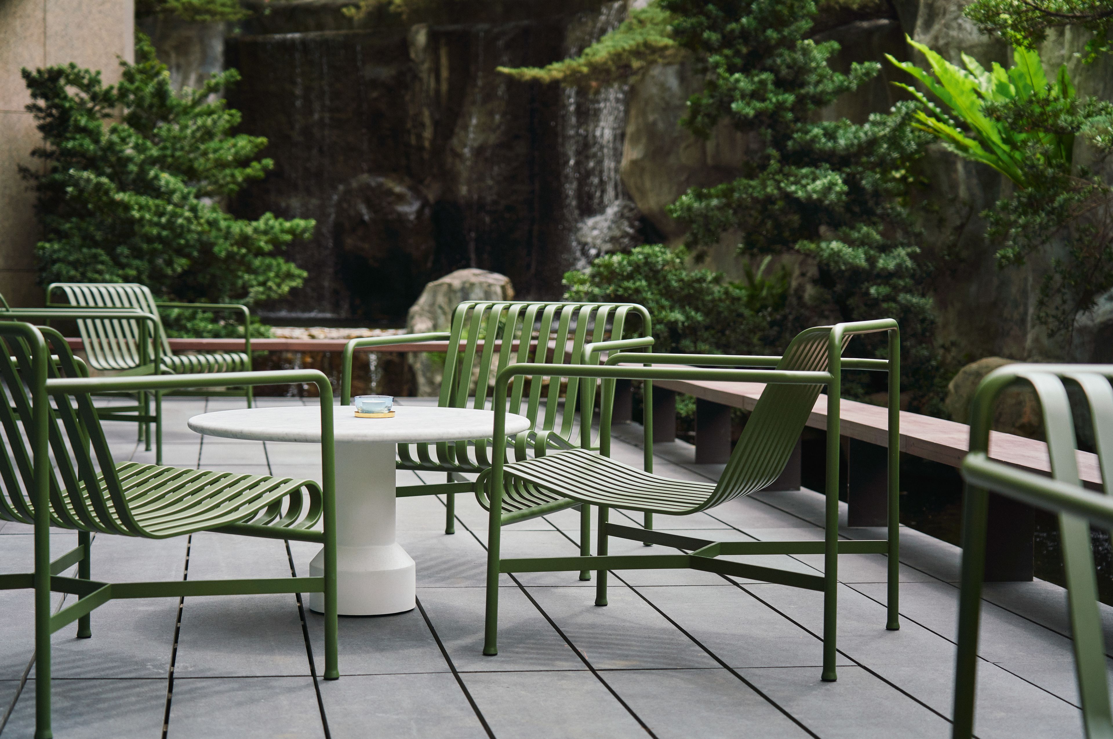 A patio area with green metal chairs and a round table, surrounded by lush greenery and a waterfall backdrop.