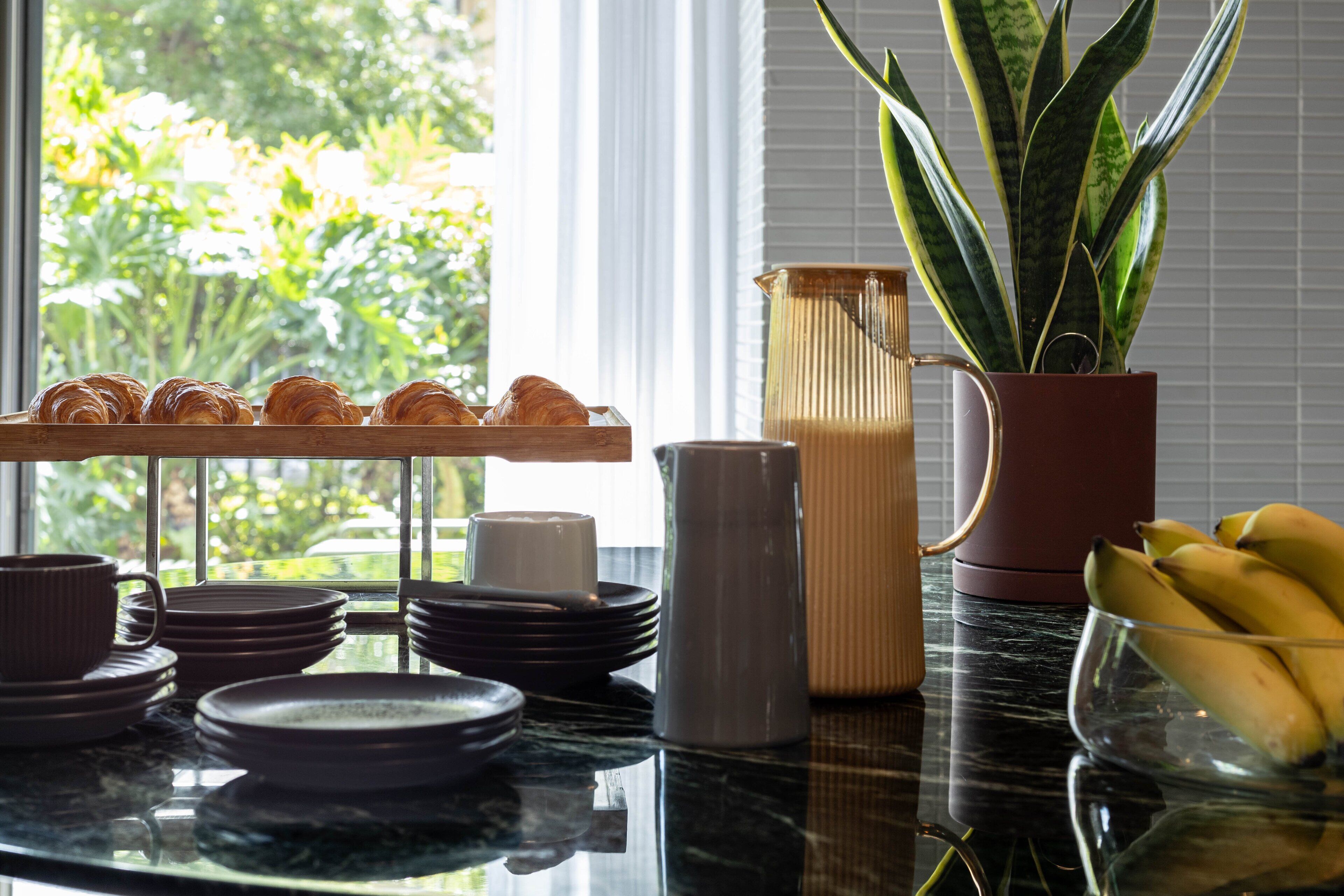 A breakfast setup with croissants, a pitcher, a potted plant, and a bowl of bananas.