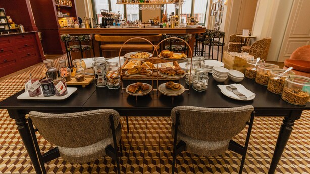 A dining area with a checkered floor, a bar in the background, and various food items on the table. - Hotel Mercier (Amsterdam)