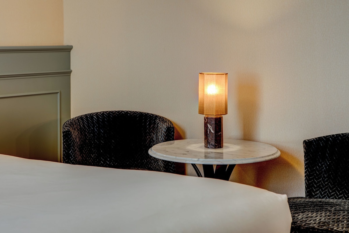 A hotel room with a round table, two upholstered chairs, a lamp, and a bed.