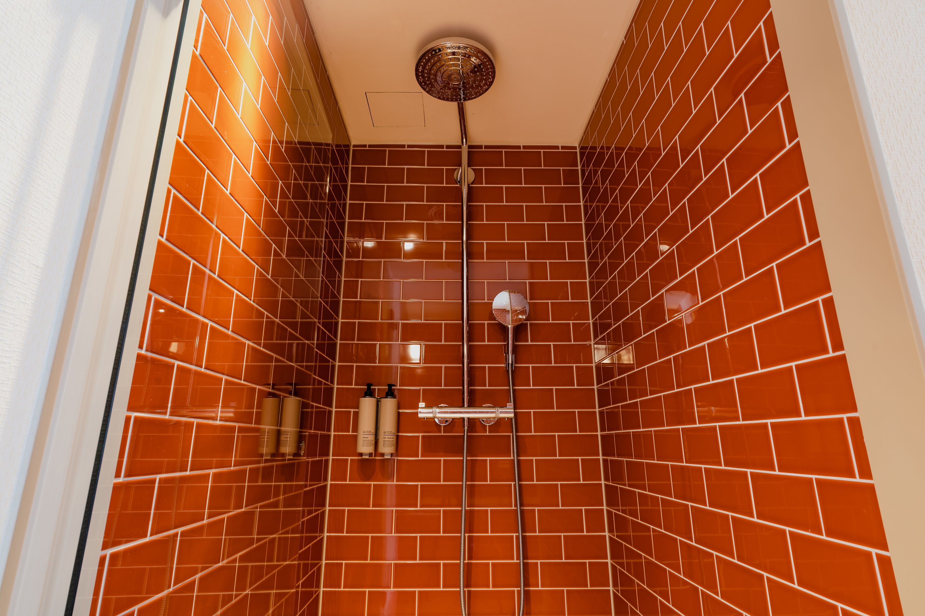 A shower with a red-tiled wall, a ceiling-mounted showerhead, and two bottles of shampoo on the wall.