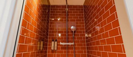 A shower with a red-tiled wall, a ceiling-mounted showerhead, and two bottles of shampoo on the wall.