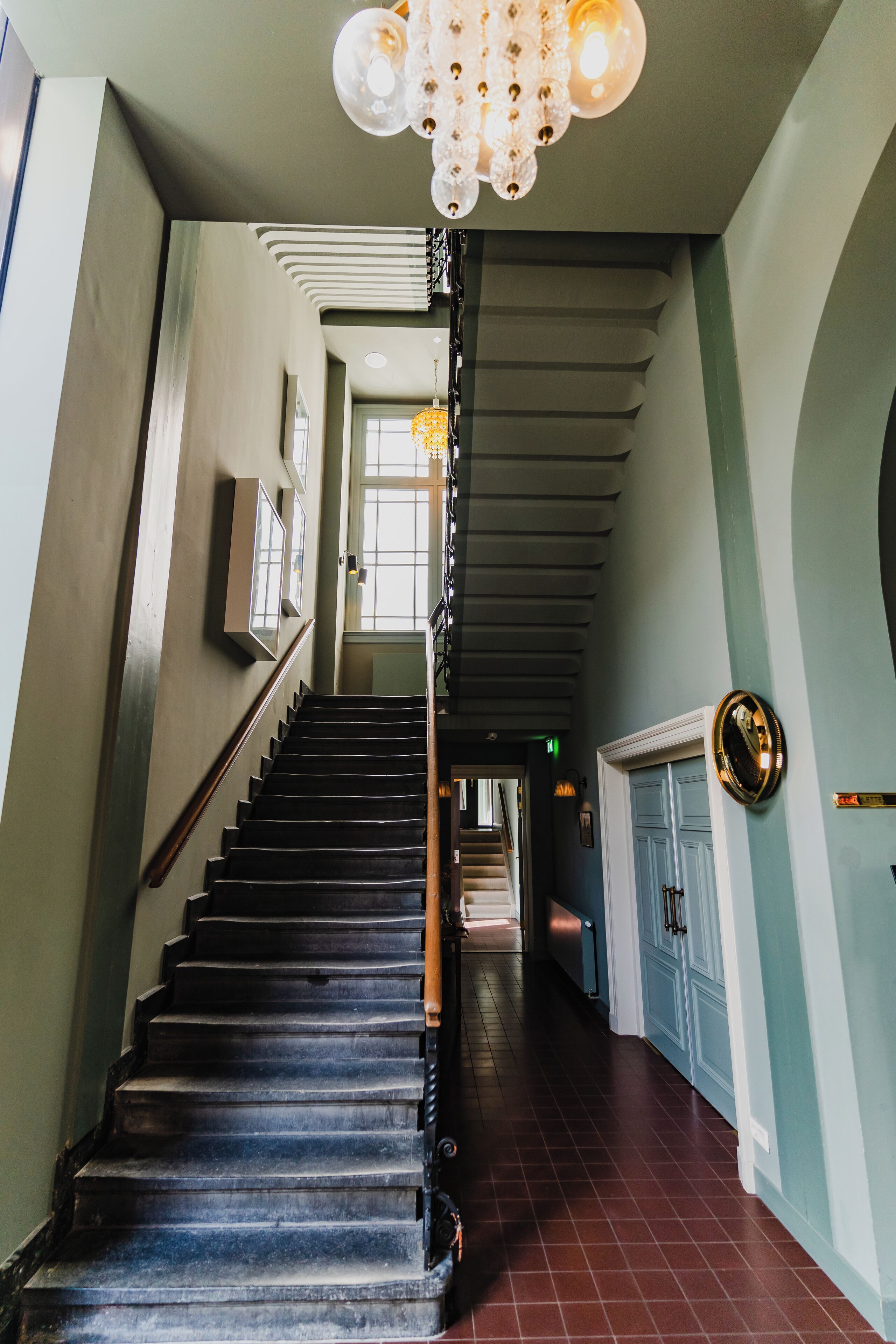 A staircase with a wooden handrail, a chandelier, and a door with a circular mirror.