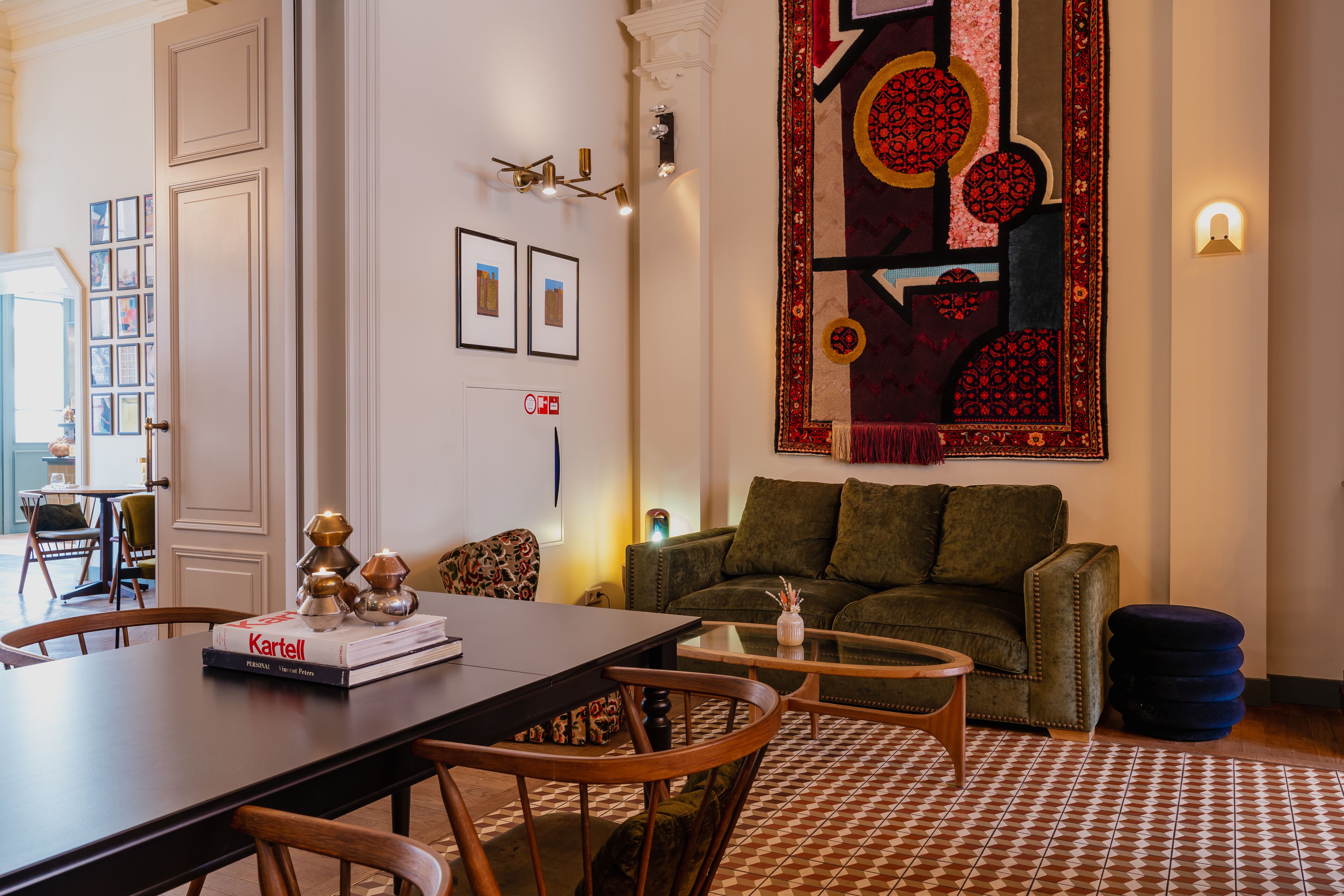 A living room with a sofa, a coffee table, and a dining table with chairs. There is a large rug on the floor and a wall with framed pictures.