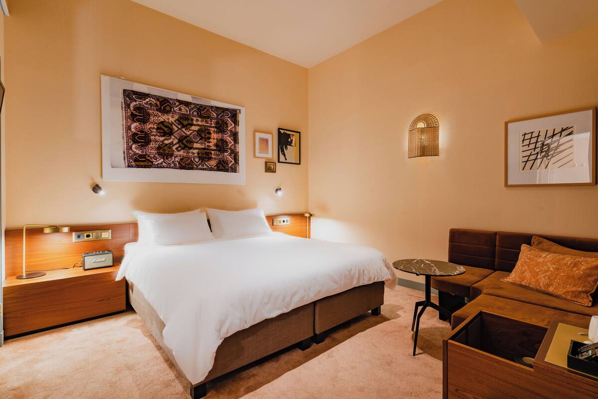 A hotel room with a large bed, a small table, a sofa, and a nightstand with a lamp.