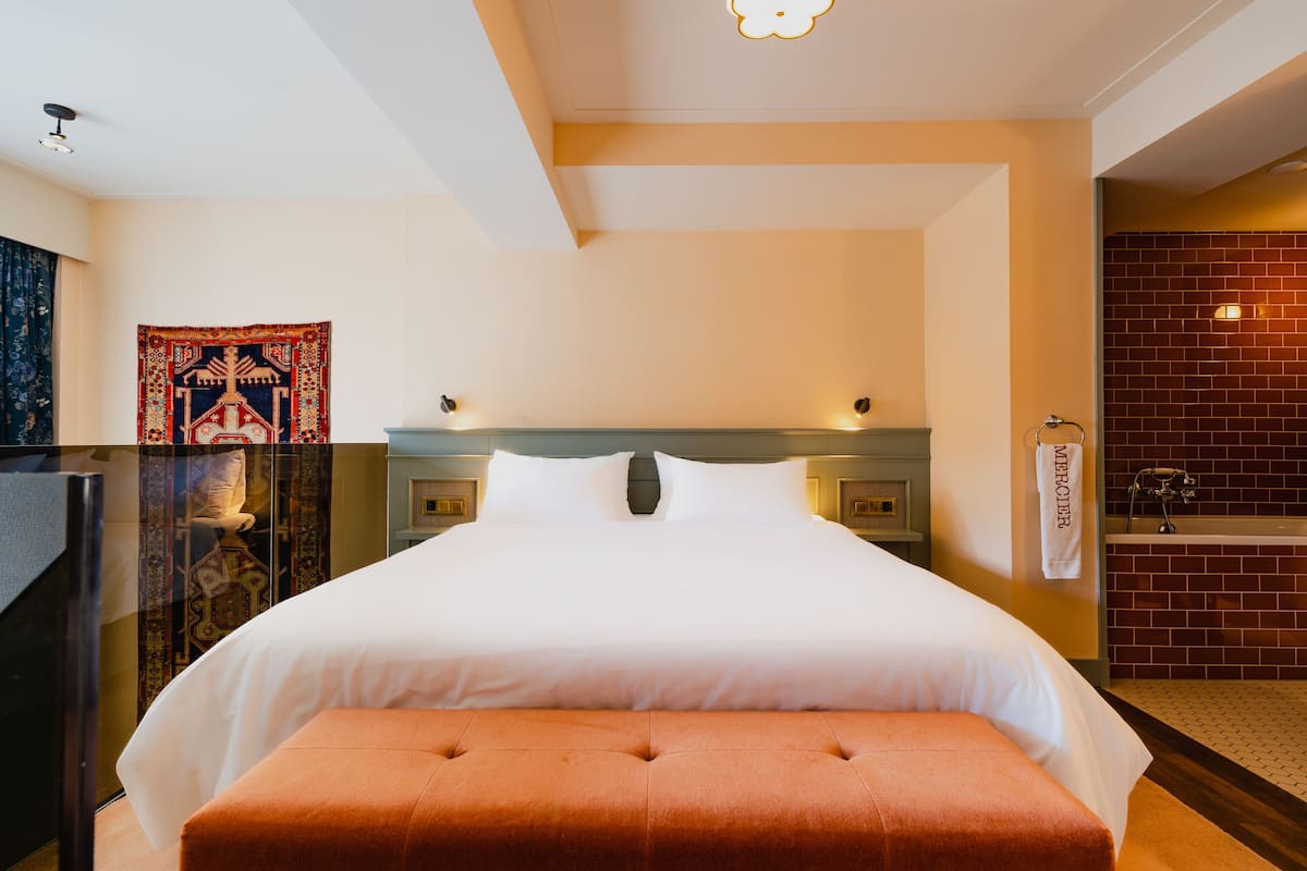 A hotel room with a large bed, a headboard, a nightstand, a bathroom with a bathtub, and a tapestry on the wall.