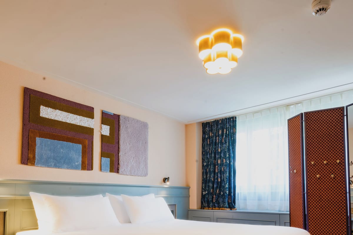 A hotel room with a large bed, a bench, a nightstand, and a window with curtains.