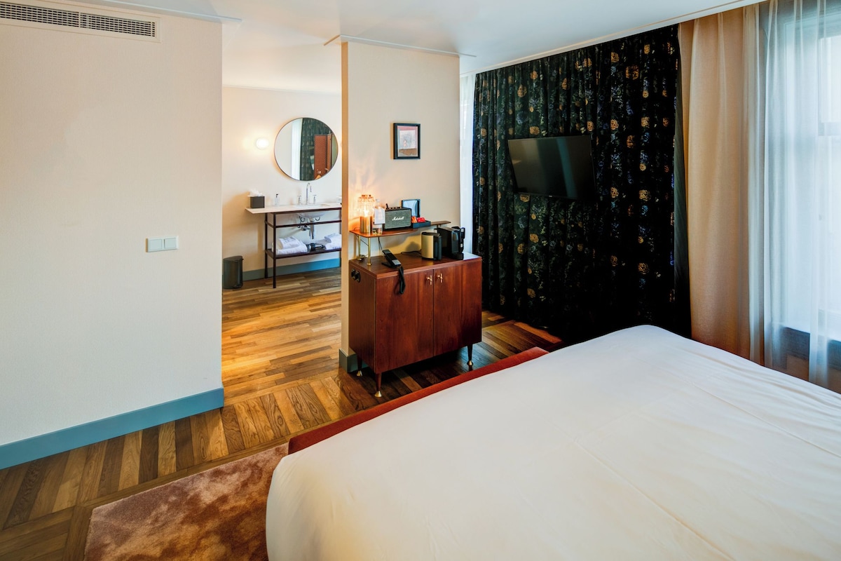 A hotel room with a large bed, a wooden dresser, a desk with a lamp, a mirror, and a TV on the wall.