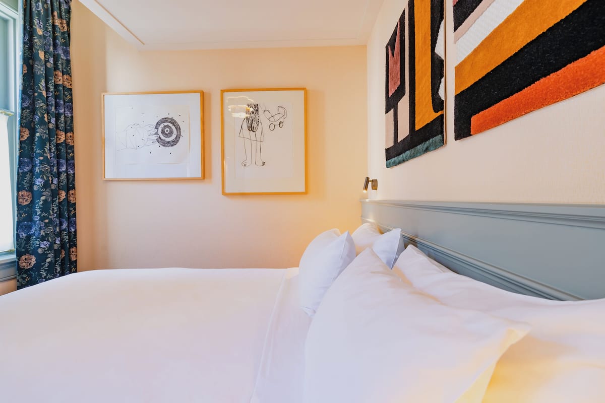 A neatly made bed with white linens in a hotel room. There are two framed artworks on the wall above the bed.