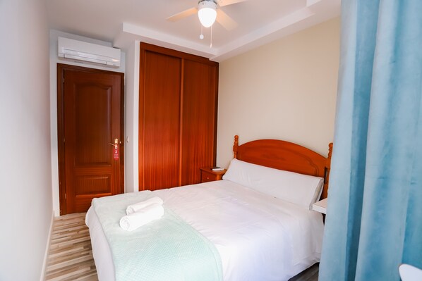 Una cama bien hecha con sábanas blancas y una manta verde, un cabecero de madera, una mesita de noche con una lámpara y un ventilador de techo.