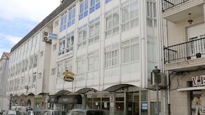 A multi-story building with large windows and balconies, featuring various storefronts and parked cars on the street. - Pension Caldas (Caldas de Reis)