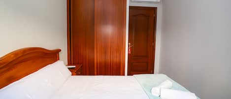 Una cama bien tendida con sábanas blancas, cabecera de madera y una manta azul claro con una toalla enrollada encima.