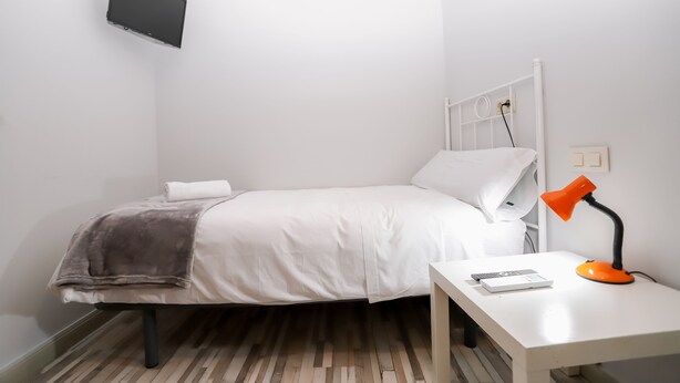 A small, neatly arranged bedroom with a bed, a bedside table with a lamp, and a TV mounted on the wall. - Pension Caldas (Caldas de Reis)