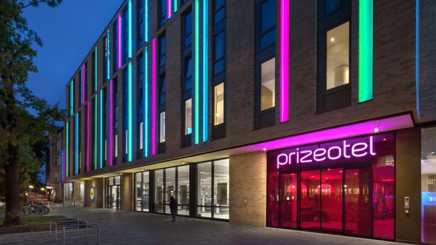 A modern building with a prominent 'prizeotel' sign, featuring a glass facade and vertical neon lights.