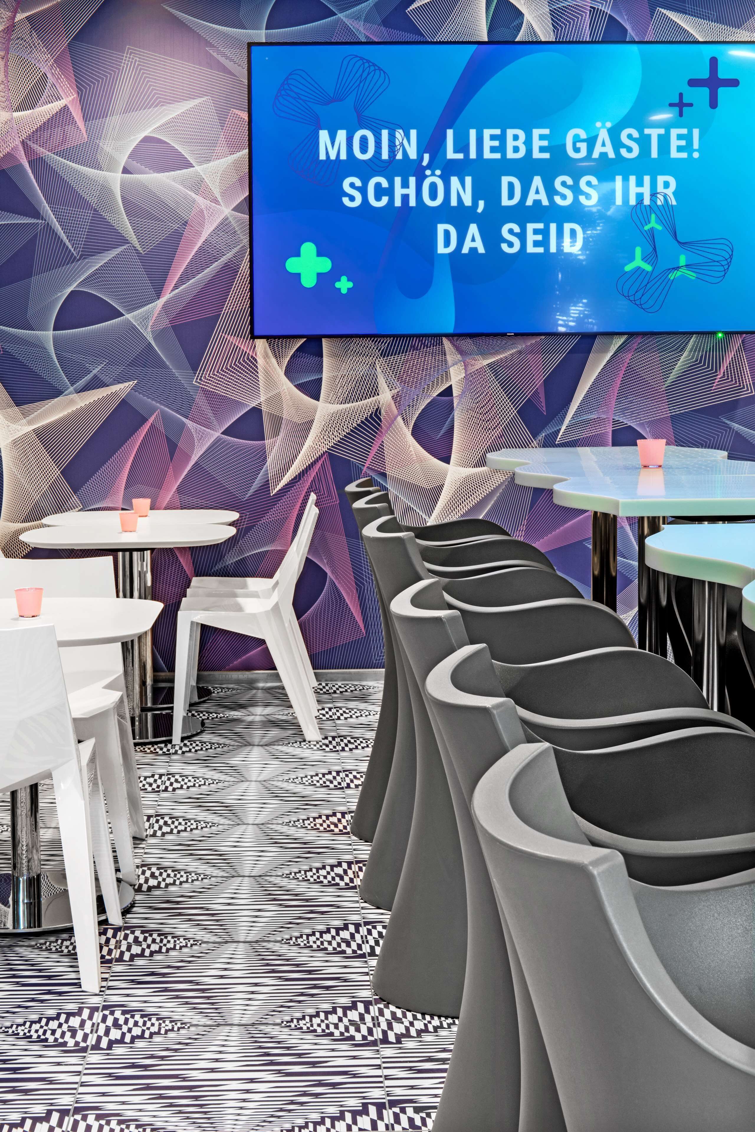 A modern restaurant interior with a geometric patterned wall, white and black chairs, and a central table with a pink cup.