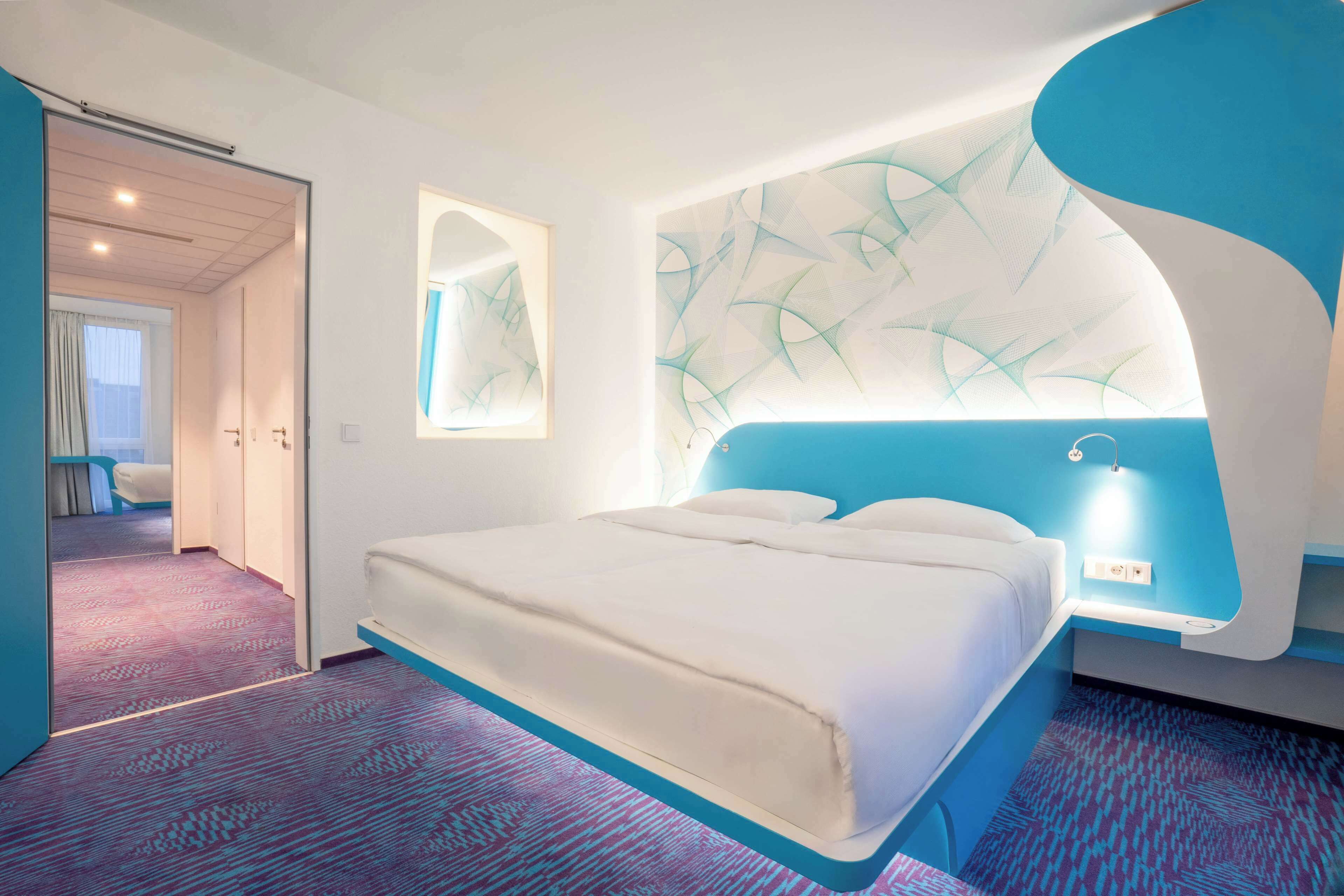 A modern hotel room with a large bed, a unique blue accent wall, and a patterned carpet.